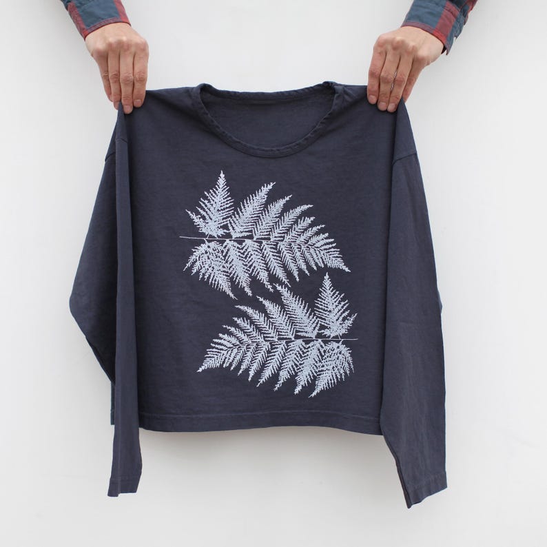May include: A navy blue long-sleeved shirt featuring a light blue fern design. The fern design is composed of two large fern fronds with detailed leaf patterns. The shirt is held up against a white background.