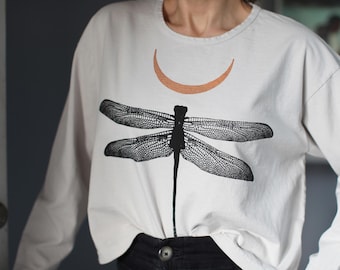May include: A long-sleeve, off-white crew neck shirt featuring a large, detailed black graphic of a dragonfly centered on the chest. Above the dragonfly, there is a smaller, metallic copper-colored crescent moon shape printed on the fabric.