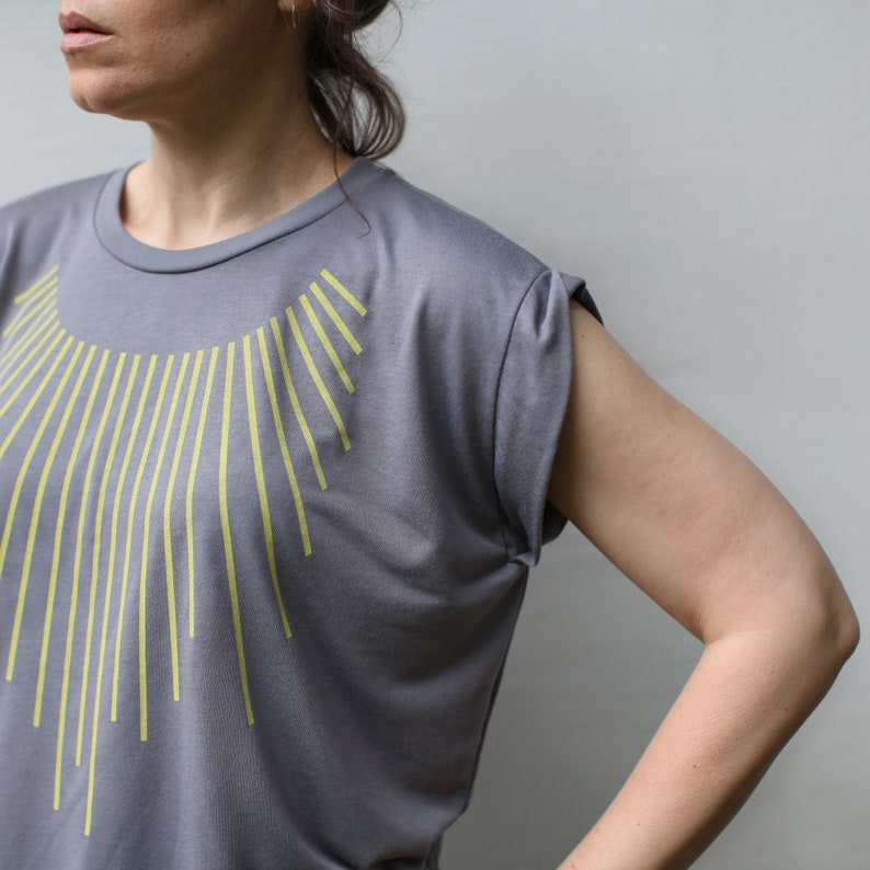 K&ouml;nnte beinhalten: Ein graues T-Shirt mit einem gelben geometrischen Design auf der Vorderseite. Das Shirt hat eine lockere Passform und kurze &Auml;rmel.