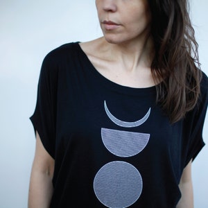 May include: Black t-shirt with a white graphic design of three stacked moons. The moons are outlined in white and have a striped pattern.