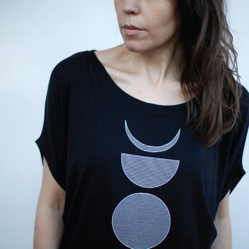 May include: Black t-shirt with a white graphic design of three stacked moons. The moons are outlined in white and have a striped pattern.