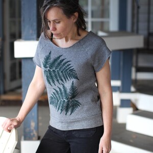 Puede incluir: Camiseta gris de manga corta con cuello redondo y un dise&ntilde;o de hojas de helecho verde oscuro en la parte delantera. La camiseta est&aacute; hecha de un material suave y se combina con pantalones negros.