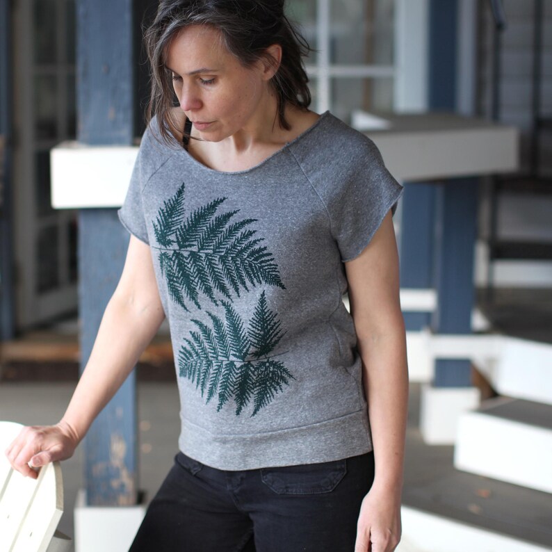 Puede incluir: Camiseta gris de manga corta con cuello redondo y un dise&ntilde;o de hojas de helecho verde oscuro en la parte delantera. La camiseta est&aacute; hecha de un material suave y se combina con pantalones negros.