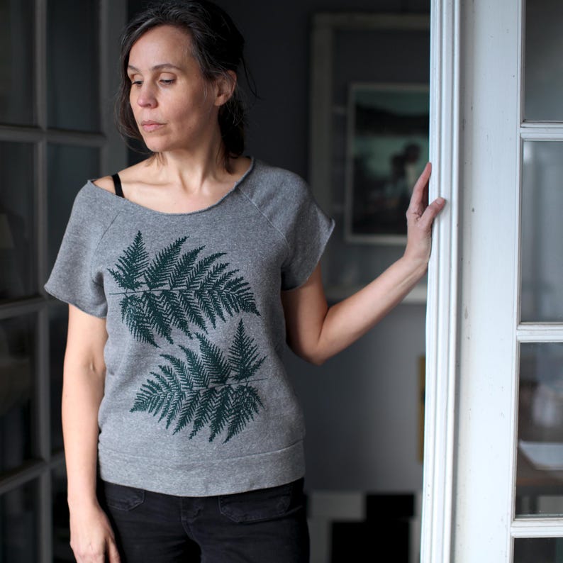 Puede incluir: Sudadera gris de manga corta con un dise&ntilde;o de hojas de helecho verde oscuro. La sudadera tiene un cuello ancho y se combina con pantalones negros. La imagen est&aacute; tomada en interiores, con una persona de pie cerca de una puerta blanca.