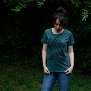Puede incluir: Una mujer lleva una camiseta verde oscuro con un estampado de helecho negro. La camiseta tiene cuello redondo y mangas cortas.