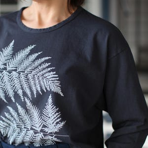 May include: A navy blue long-sleeve shirt featuring a white fern leaf design on the front. The shirt has a crew neck and a relaxed fit. The fern design is detailed and covers a portion of the shirt's front.