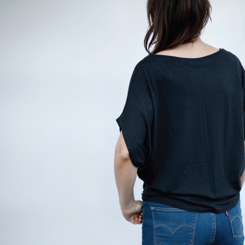 May include: A black, loose-fitting, short-sleeved shirt with a boat neck. The shirt is tucked into a pair of blue jeans.