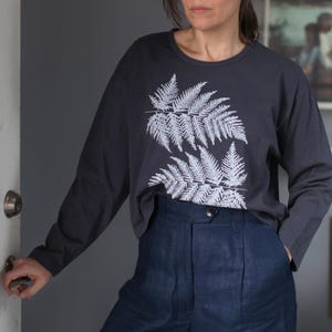 May include: Dark gray long-sleeve shirt with a white fern leaf design. The shirt is paired with high-waisted, dark blue pants. The outfit is casual and comfortable, suitable for everyday wear.