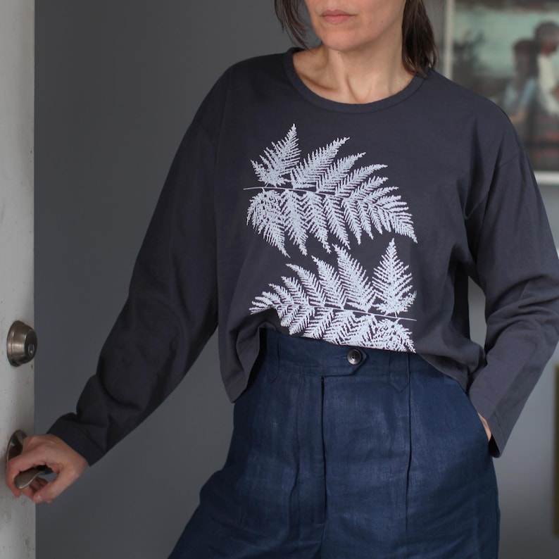 May include: Dark gray long-sleeve shirt with a white fern leaf design. The shirt is paired with high-waisted, dark blue pants. The outfit is casual and comfortable, suitable for everyday wear.
