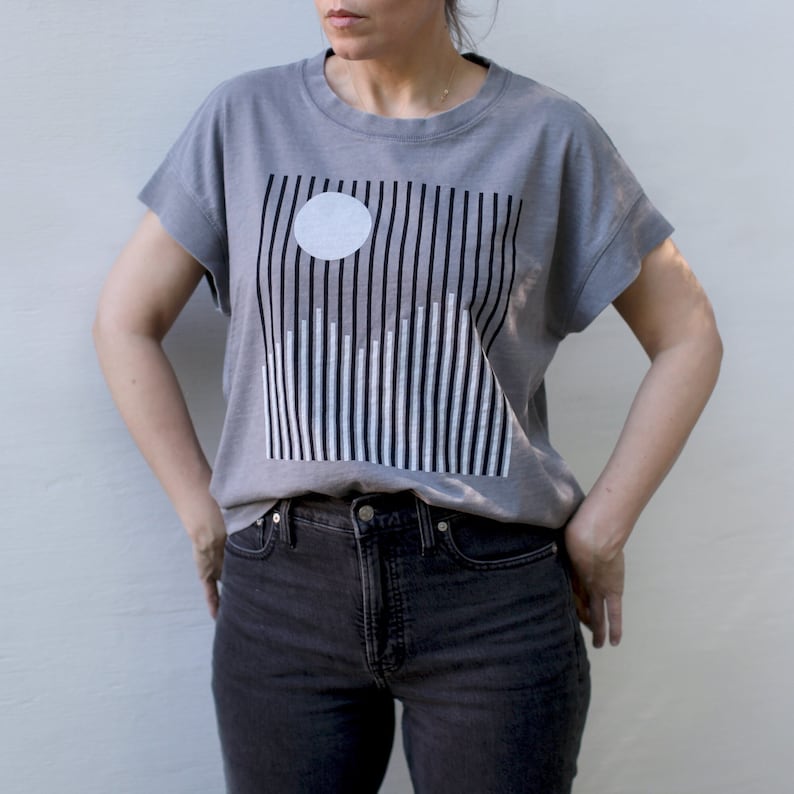 May include: A gray boxy top with black and white graphic print of a moon over snowy mountains. The shirt has cap sleeves and a round neck.