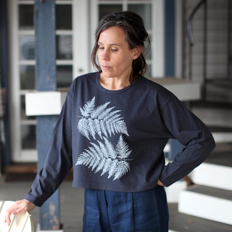 May include: A navy blue long-sleeve shirt with a white fern leaf design. The shirt is a cropped style and is paired with navy blue pants. The model is standing outside near a white door and a blue post.