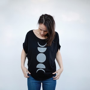 May include: A woman wearing a black t-shirt with a white moon phases design. The shirt has a relaxed fit and a scoop neck.