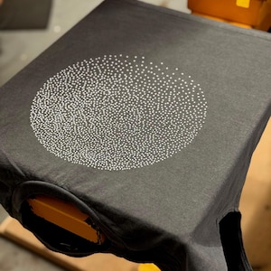 Op de afbeelding: Een donkergrijs T-shirt met een wit cirkelontwerp gemaakt van kleine stippen.