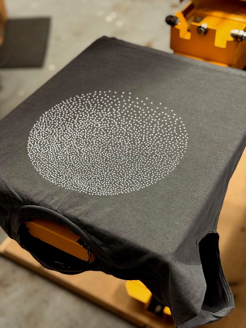 Op de afbeelding: Een donkergrijs T-shirt met een wit cirkelontwerp gemaakt van kleine stippen.