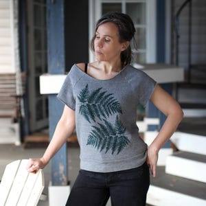 Puede incluir: Sudadera gris de manga corta con un dise&ntilde;o de hojas de helecho verde oscuro. La sudadera tiene un corte holgado y un escote sin rematar. La modelo lleva pantalones negros y est&aacute; de pie en un porche.
