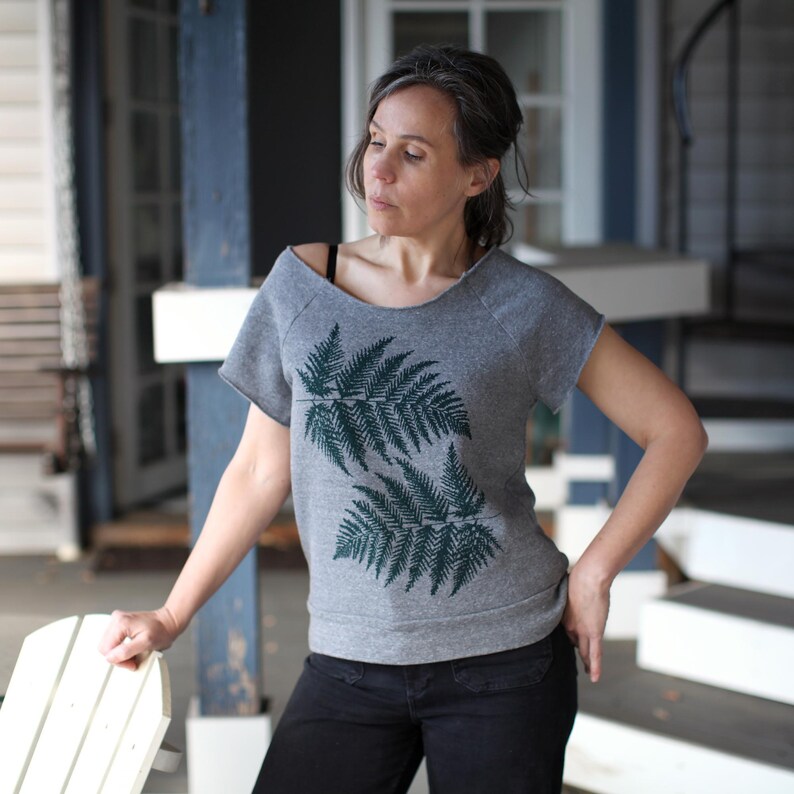 Puede incluir: Sudadera gris de manga corta con un dise&ntilde;o de hojas de helecho verde oscuro. La sudadera tiene un corte holgado y un escote sin rematar. La modelo lleva pantalones negros y est&aacute; de pie en un porche.