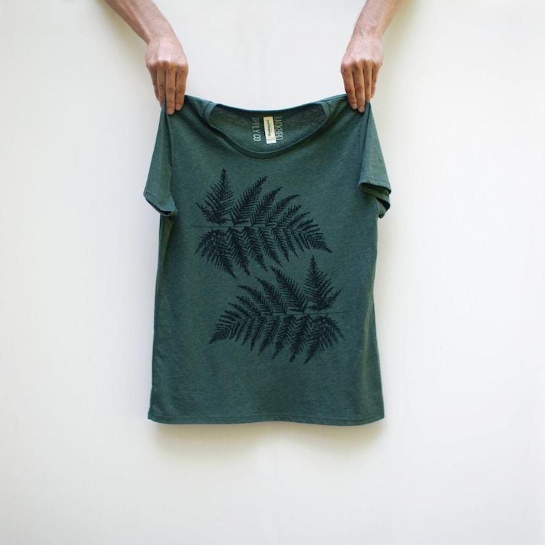 Puede incluir: Una camiseta verde con un estampado de helecho negro. La camiseta tiene cuello redondo y mangas cortas.