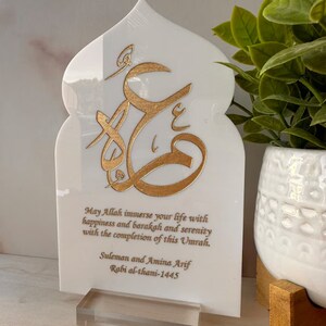 Puede incluir: Placa de m&aacute;rmol blanco con un dise&ntilde;o de caligraf&iacute;a &aacute;rabe dorado y un mensaje que dice "Que Allah inunde tu vida de felicidad, barakah y serenidad con la finalizaci&oacute;n de esta Umrah. Suleman y Amina Arif Rabi al-thani-1445".