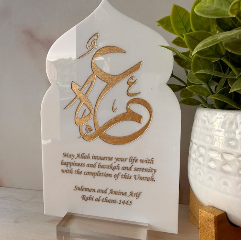 Puede incluir: Placa de m&aacute;rmol blanco con un dise&ntilde;o de caligraf&iacute;a &aacute;rabe dorado y un mensaje que dice "Que Allah inunde tu vida de felicidad, barakah y serenidad con la finalizaci&oacute;n de esta Umrah. Suleman y Amina Arif Rabi al-thani-1445".