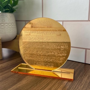 May include: A gold-colored round acrylic plaque with Arabic calligraphy etched into the surface. The plaque is mounted on a matching gold-colored stand.