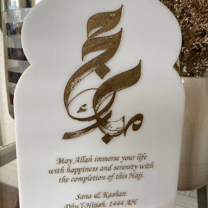 Puede incluir: Placa blanca con un dise&ntilde;o de caligraf&iacute;a &aacute;rabe dorado. La placa tiene un mensaje que dice: "Que Allah inunde tu vida de felicidad y serenidad con la finalizaci&oacute;n de este Hajj. Sana & Kashan Dhu'l-Hijjah, 1444 AH".