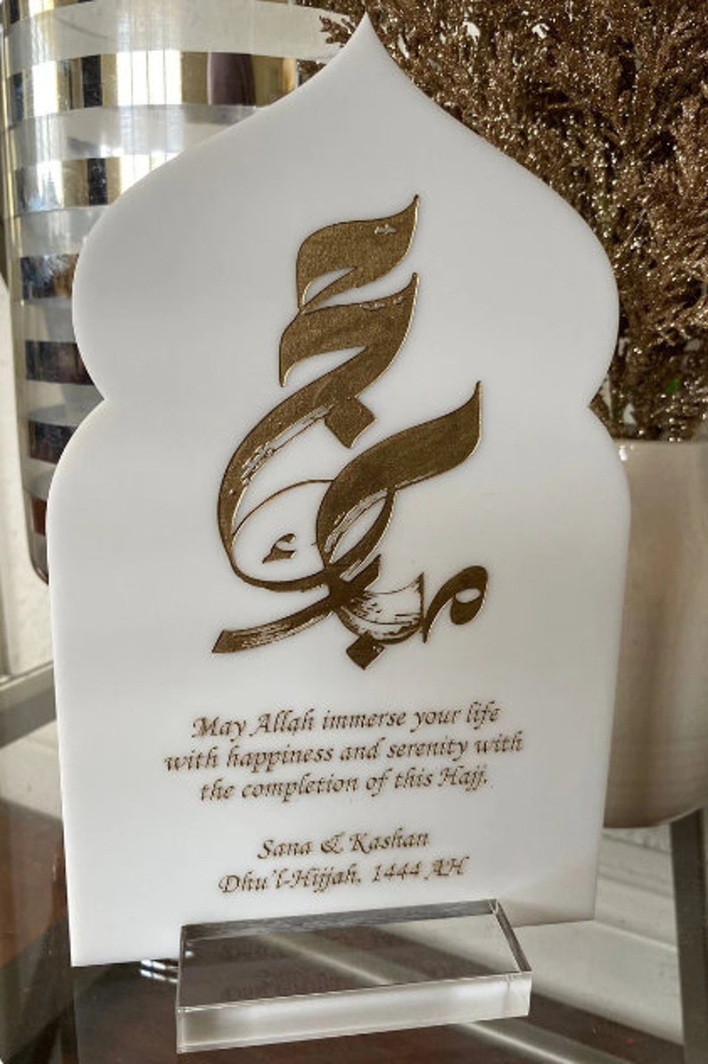 Puede incluir: Placa blanca con un dise&ntilde;o de caligraf&iacute;a &aacute;rabe dorado. La placa tiene un mensaje que dice: "Que Allah inunde tu vida de felicidad y serenidad con la finalizaci&oacute;n de este Hajj. Sana & Kashan Dhu'l-Hijjah, 1444 AH".