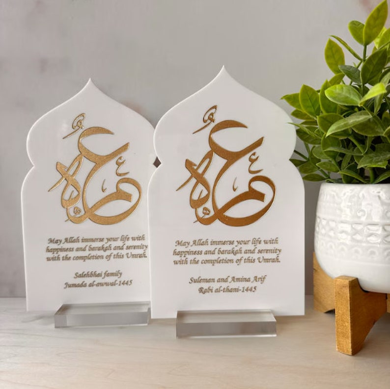 Puede incluir: Dos placas decorativas blancas con caligraf&iacute;a dorada y un mensaje sobre la felicidad y la serenidad. Las placas tienen forma de arco y se colocan sobre bases de acr&iacute;lico transparente. El texto en las placas dice: "Que Allah inunde tu vida de felicidad, barakah y serenidad con la finalizaci&oacute;n de esta Umrah." La primera placa tiene el texto "Salehbhai family Jumada al-awwal-1445" y la segunda placa tiene el texto "Suleman y Amina Arif Rabi al-thani-1445".