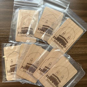 Könnte beinhalten: Eine Sammlung rechteckiger Holzplaketten, jede mit einer detaillierten Gravur einer religiösen Struktur, darunter ein zentrales kubusförmiges Gebäude und zwei Minarette. Die Plaketten sind in transparenten Plastiktüten verpackt.