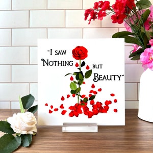 May include: A white square sign with a red rose and the text "I saw nothing but beauty". The sign is on a clear acrylic stand.