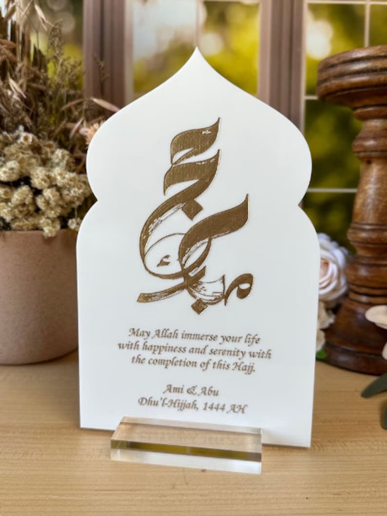 Puede incluir: Tarjeta blanca con un dise&ntilde;o de caligraf&iacute;a &aacute;rabe dorada y un mensaje que dice "Que Allah inunde tu vida de felicidad y serenidad con la finalizaci&oacute;n de este Hajj. Ami & Abu Dhu'l-Hijjah, 1444 AH".