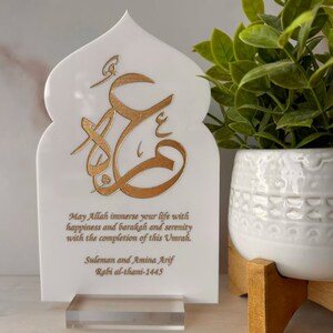 Puede incluir: Placa decorativa blanca con un dise&ntilde;o de caligraf&iacute;a &aacute;rabe dorado y un mensaje que dice "Que Allah inunde tu vida de felicidad, barakah y serenidad con la finalizaci&oacute;n de esta Umrah. Suleman y Amina Arif Rabi al-thani-1445".