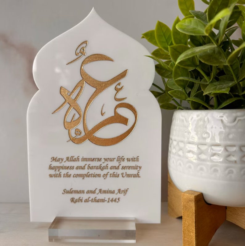 Puede incluir: Placa decorativa blanca con un dise&ntilde;o de caligraf&iacute;a &aacute;rabe dorado y un mensaje que dice "Que Allah inunde tu vida de felicidad, barakah y serenidad con la finalizaci&oacute;n de esta Umrah. Suleman y Amina Arif Rabi al-thani-1445".