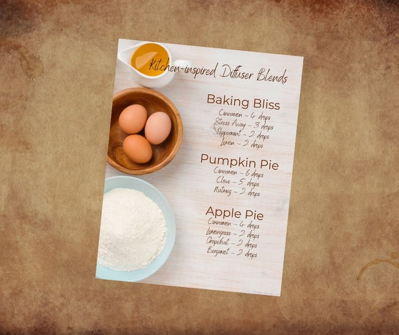 May include: A white card with brown text lists kitchen-inspired diffuser blends with essential oil recipes for Baking Bliss, Pumpkin Pie, and Apple Pie. The card is on a wooden surface with a bowl of flour and a bowl of eggs.
