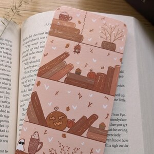Könnte beinhalten: Ein rosa-braunes Lesezeichen mit Herbstthema. Das Lesezeichen zeigt einen Stapel Bücher, einen Kürbis, ein Gespenst, eine Topfpflanze und eine Tasse Kaffee. Das Lesezeichen ist perfekt für Bücherwürmer und alle, die die Herbstsaison lieben.