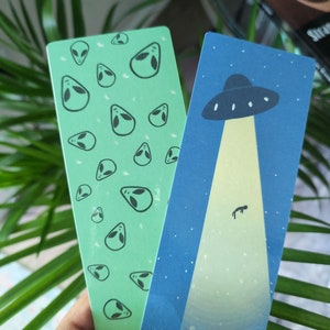 Puede incluir: Dos marcapáginas con temática extraterrestre. Un marcapáginas es verde con un patrón repetido de cabezas de extraterrestres. El otro marcapáginas es azul con un OVNI y una persona siendo abducida por un rayo de luz.