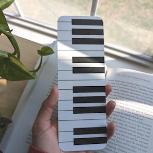 May include: A white bookmark featuring a black piano keyboard design. The bookmark is being held in a hand.
