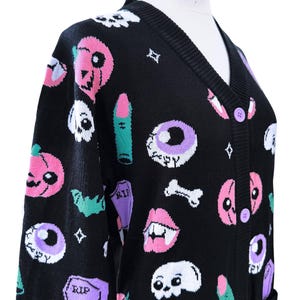 May include: A black knit cardigan featuring a repeating pattern of pink pumpkins, white skulls, purple eyeballs, green fingers, white bones, and purple tombstones with the text RIP. The garment has a V-neckline and light purple buttons down the front.