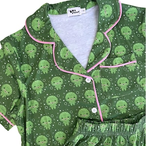 May include: A dark green pajama set featuring a repeating pattern of small cartoon swamp creatures and white bubbles. The top has a collared neckline, short sleeves, a chest pocket, and white buttons, all accented with thin pink piping trim.