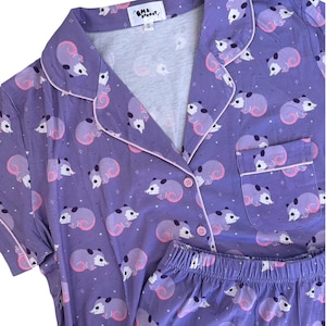 May include: A purple pajama set featuring a repeating pattern of small white and gray opossums with pink tails and ears. The short-sleeved button-down top has a collar, a chest pocket, and pink piping. The label reads EM & SPROUT and size S.