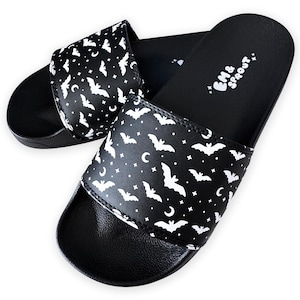 May include: Black slide sandals with a white bat and crescent moon pattern. The sandals have a black sole and a wide strap across the top. The text "EM & Sprout" is printed in white on the footbed.