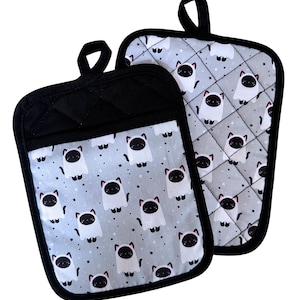 May include: Two gray and black oven mitts with a cat ghost pattern. The mitts have a black quilted pocket and trim, and a black loop for hanging. The cat ghosts are white with black faces and pink ears, set against a gray background with stars.
