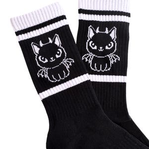 May include: Pair of black socks with white stripes and a white cartoon cat graphic with bat wings. The socks have a ribbed cuff and are made of a soft material.