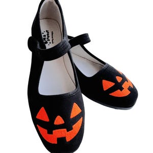 Puede incluir: Zapatillas de lona negras con caras de calabaza naranja. Las zapatillas tienen una correa en la parte superior y están diseñadas para Halloween.