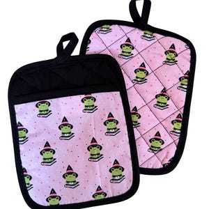 May include: Two quilted pot holders with black trim and hanging loops. The main fabric is light pink with a repeating pattern of green frog illustrations wearing witch hats and sitting on books. The pot holders are designed for kitchen use.
