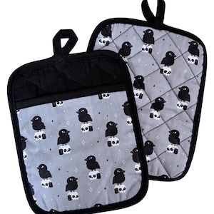 May include: Two black and gray oven mitts. The gray fabric is patterned with black crows perched on white skulls. The mitts have black quilted backs and black trim, with a loop for hanging.