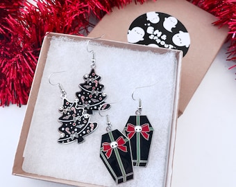 May include: A pair of earrings in a gift box. One pair is shaped like black Christmas trees with bats, candy canes, and skulls. The other pair is coffin-shaped, black with red bows and skull accents. The box has a sticker with ghost illustrations.