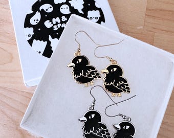 May include: Black bird-shaped earrings with gold and silver hooks, displayed in a white box. The earrings have a detailed design with a black body and white accents. The box has a black and white graphic of ghosts and the text "EM & SPA".