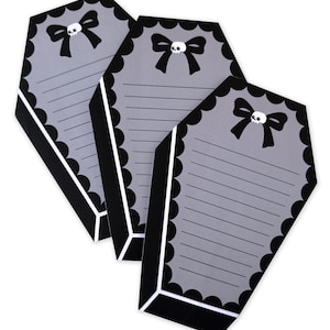 May include: Three coffin-shaped notepads with gray backgrounds and black scalloped edges. Each notepad features a black bow with a skull in the center and horizontal lines for writing. The notepads have a white border.