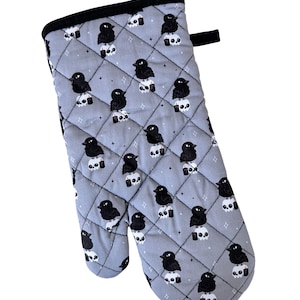 May include: A gray quilted oven mitt featuring a repeating pattern of black crows perched on white skulls. The mitt has a black trim and a hanging loop. The design includes small star accents.