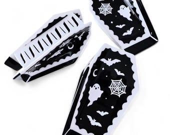 Puede incluir: Tres platos para baratijas en forma de ataúd en blanco y negro. Cada plato presenta un borde festoneado y está decorado con murciélagos, una telaraña, un fantasma y una luna creciente. Los platos están dispuestos sobre un fondo blanco.